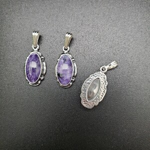 Peut inclure: Trois pendentifs en argent avec des pierres de forme ovale. Deux pendentifs pr&eacute;sentent des pierres violettes et blanches, tandis que le troisi&egrave;me pendentif a une pierre argent&eacute;e.
