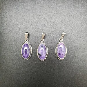 Peut inclure: Trois pendentifs en argent avec des pierres ovales de couleur violette et blanche. Les pendentifs sont sur un fond gris fonc&eacute;.