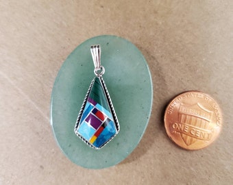 May include: A silver pendant with a geometric design featuring turquoise, red, purple, and yellow stones. The pendant is approximately the size of a US penny.