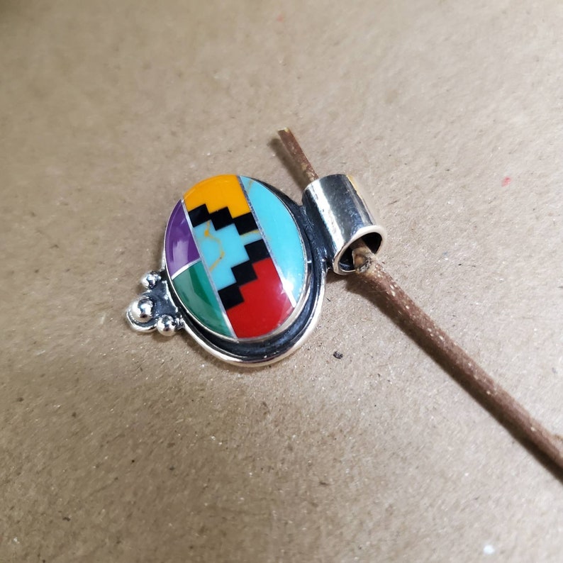 May include: A silver pendant with a colorful inlay of turquoise, red, yellow, green, purple, and black stones. The pendant is oval shaped and has a bail for attaching a chain.