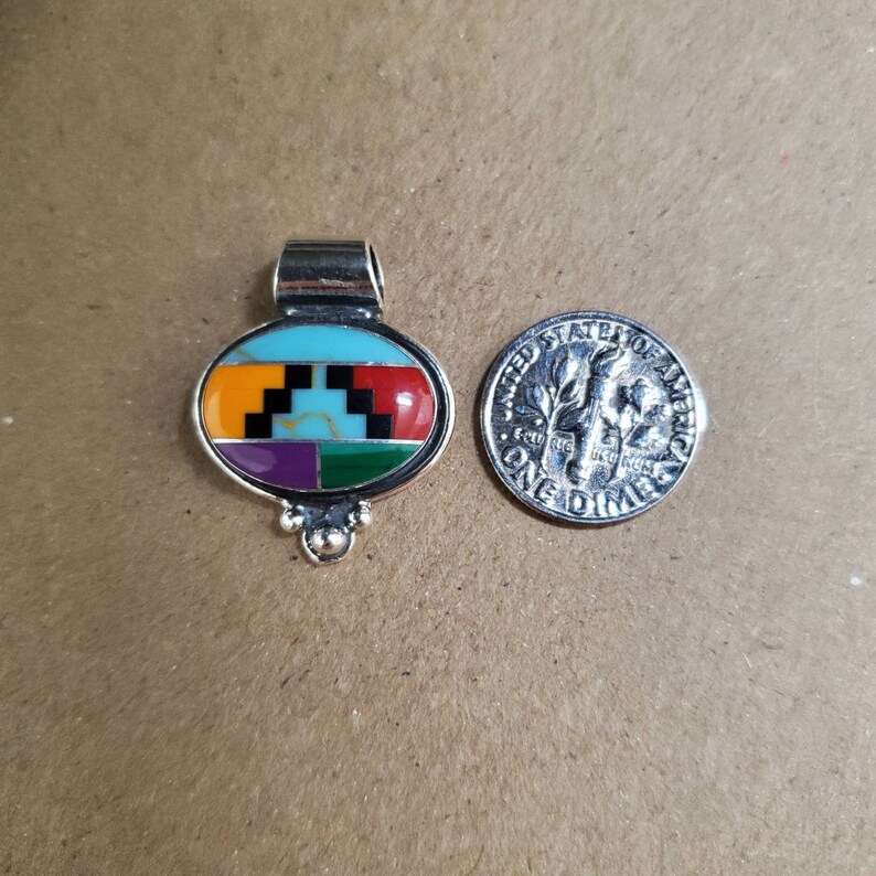 May include: A silver pendant with an oval-shaped turquoise, red, orange, purple, green, and black inlay. The pendant is about the size of a dime.