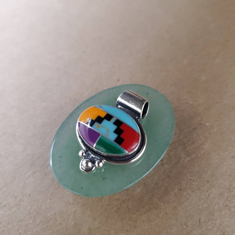 May include: A silver pendant with a colorful inlay of turquoise, red, yellow, green, purple, and black stones. The pendant is oval shaped and has a bail for attaching a chain.