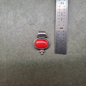 K&ouml;nnte beinhalten: Ein silberner Anh&auml;nger mit einem roten Korallen-Stein. Der Anh&auml;nger hat einen Durchmesser von etwa 2,5 cm.
