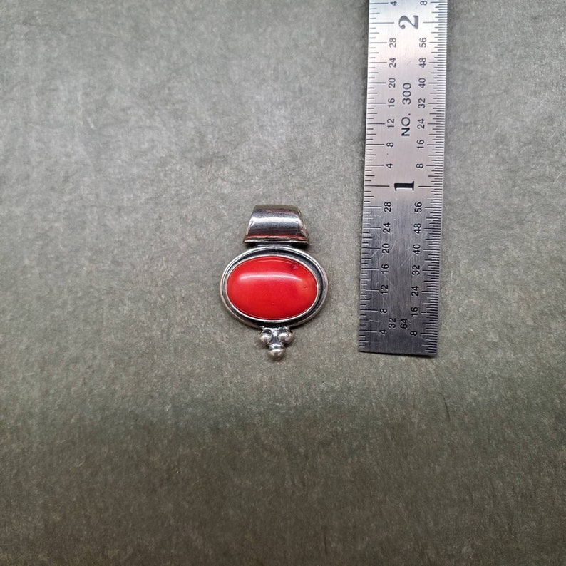 K&ouml;nnte beinhalten: Ein silberner Anh&auml;nger mit einem roten Korallen-Stein. Der Anh&auml;nger hat einen Durchmesser von etwa 2,5 cm.