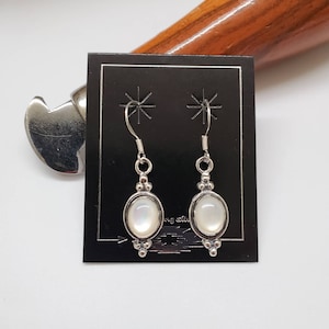 May include: A pair of silver earrings with oval-shaped white mother-of-pearl stones. The earrings have a simple design with a small cluster of beads at the bottom.