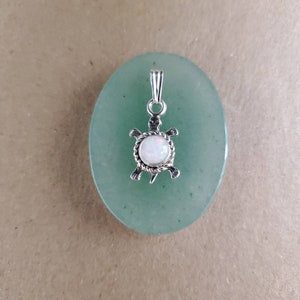Puede incluir: Un colgante de plata en forma de tortuga con una piedra de ópalo blanco. El colgante está sobre una piedra de jade verde.