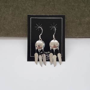May include: Silver earrings with a round, silver charm featuring a floral design. The charms are attached to three black feathers.