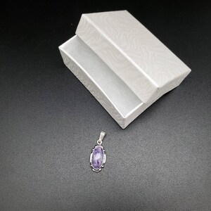 Peut inclure: Un pendentif en argent avec une pierre ovale violette sertie dans un cadre en argent. Le pendentif est sur un fond noir.