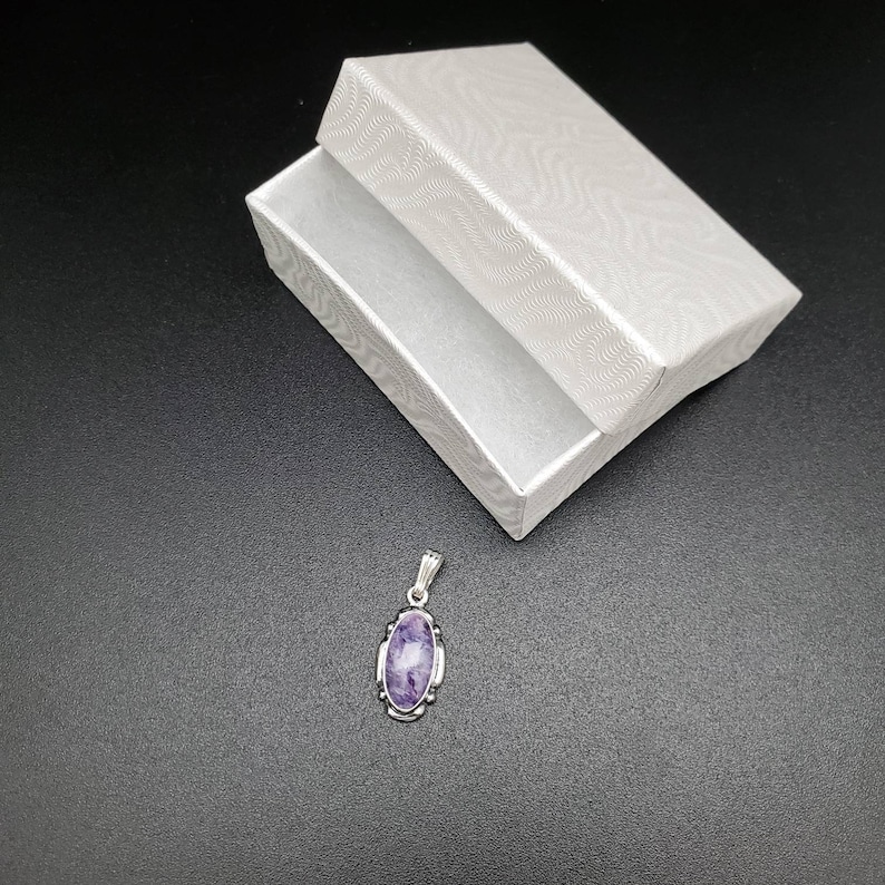 Peut inclure: Un pendentif en argent avec une pierre ovale violette sertie dans un cadre en argent. Le pendentif est sur un fond noir.