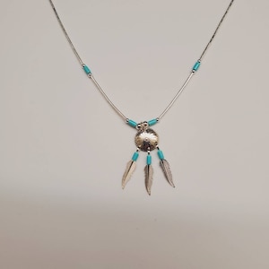 May include: A silver necklace with a turquoise pendant featuring a circular design and three feathers. The necklace is adorned with turquoise beads.
