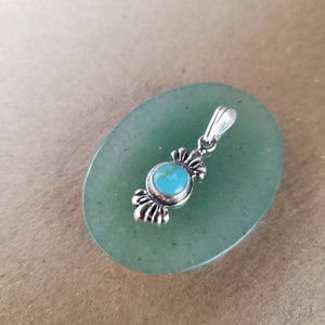 Puede incluir: Un colgante de plata con una piedra turquesa engastada en un marco de plata decorativo. El colgante está sobre una superficie de piedra verde.