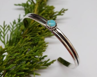 May include: A polished silver cuff bracelet featuring a turquoise gemstone. The bracelet has a simple, elegant design with a round, light blue turquoise stone set in a decorative bezel. The bracelet is approximately 16 cm in circumference.