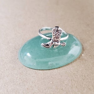 May include: A silver ring featuring a detailed cowboy boot design. The ring is displayed on a light green stone.