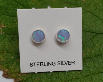 Könnte beinhalten: Ein Paar Ohrstecker aus Sterlingsilber mit runden Opal-Steinen. Die Opale sind hellblau mit irisierenden grün- und blautönen.