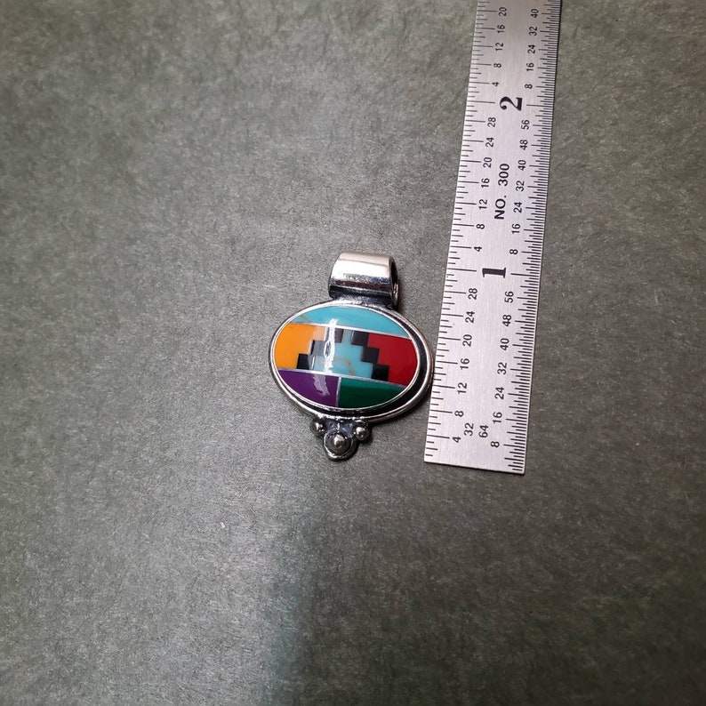 May include: A silver pendant with an oval-shaped inlay of turquoise, red, orange, purple, green, and black stones. The pendant is approximately 1 inch in length.