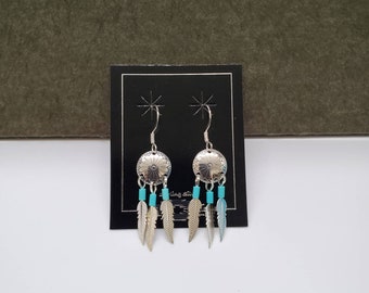 May include: Silver dangle earrings with turquoise beads and silver feathers. The earrings are displayed on a black card with a white star design.