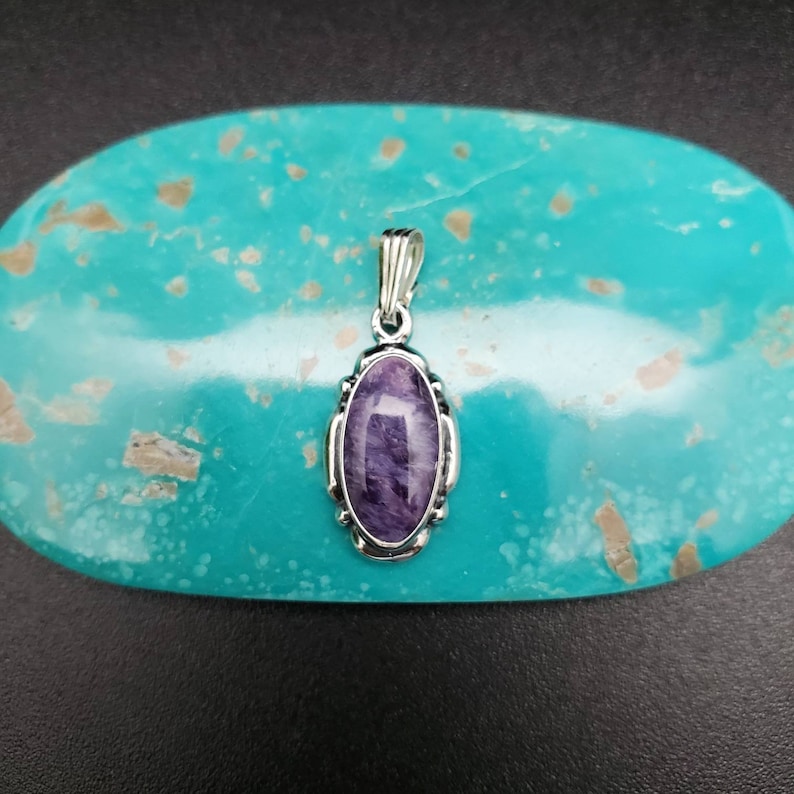 Peut inclure: Un pendentif en argent avec une pierre ovale de couleur violette et blanche sertie dans une b&eacute;li&egrave;re en argent. Le pendentif est sur un fond turquoise.