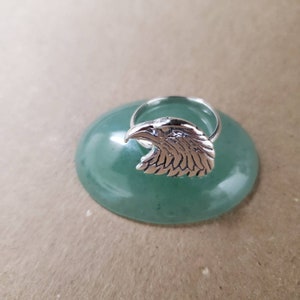May include: A silver ring with an eagle head design. The ring is displayed on a green stone surface.