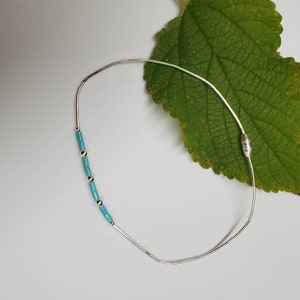 Op de afbeelding: Een zilveren armband met turquoise blauwe kralen. De armband ligt op een witte achtergrond met een groen blad.