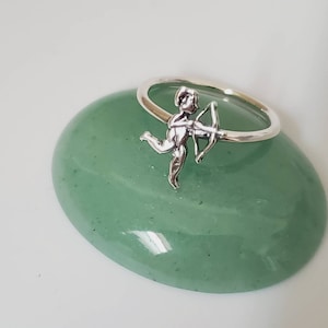 Könnte beinhalten: Ein silberner Ring mit einem Amor-Motiv. Der Amor hält einen Bogen und Pfeil und rennt nach rechts. Der Ring liegt auf einer grünen Steinoberfläche.