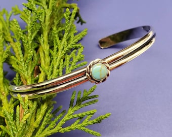 May include: A silver bangle bracelet featuring a turquoise stone set in a gold-toned bezel. The bracelet is double-layered and has a simple, elegant design.