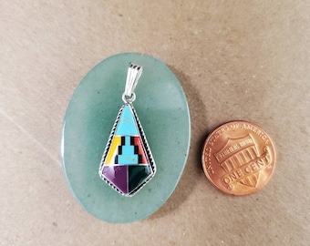 May include: A silver pendant with a geometric design featuring turquoise, black, red, yellow, purple, and green inlays. The pendant is displayed on a light green oval stone next to a one-cent coin.