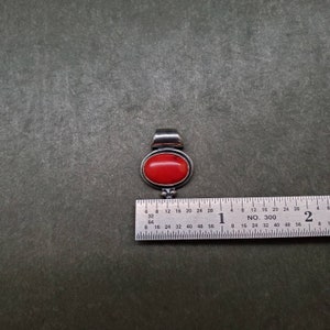 K&ouml;nnte beinhalten: Ein silberner Anh&auml;nger mit einem roten ovalen Edelstein. Der Anh&auml;nger ist etwa 2,5 cm lang und hat ein schlichtes Design.