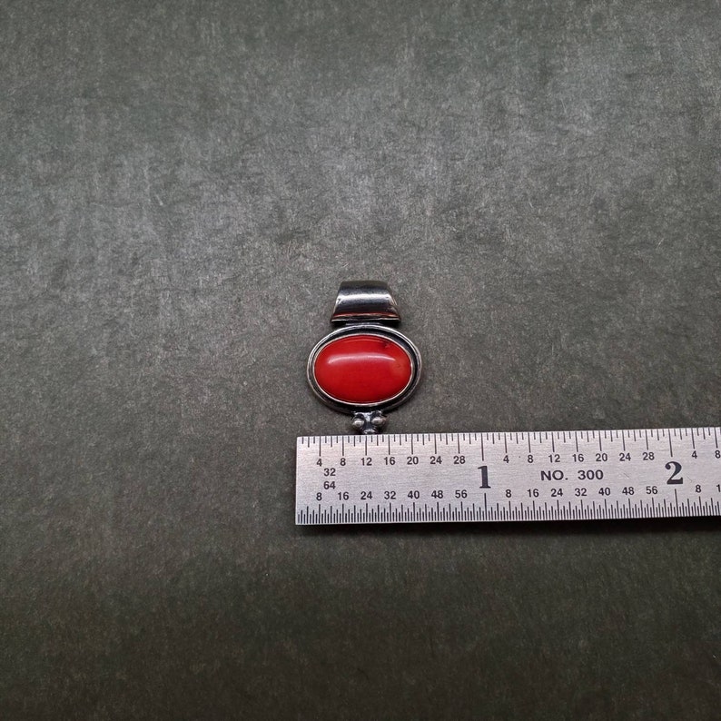 K&ouml;nnte beinhalten: Ein silberner Anh&auml;nger mit einem roten ovalen Edelstein. Der Anh&auml;nger ist etwa 2,5 cm lang und hat ein schlichtes Design.