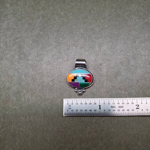 May include: A silver pendant with an oval-shaped inlay of turquoise, red, green, purple, and black stones. The pendant has a silver bail and three small silver beads at the bottom.