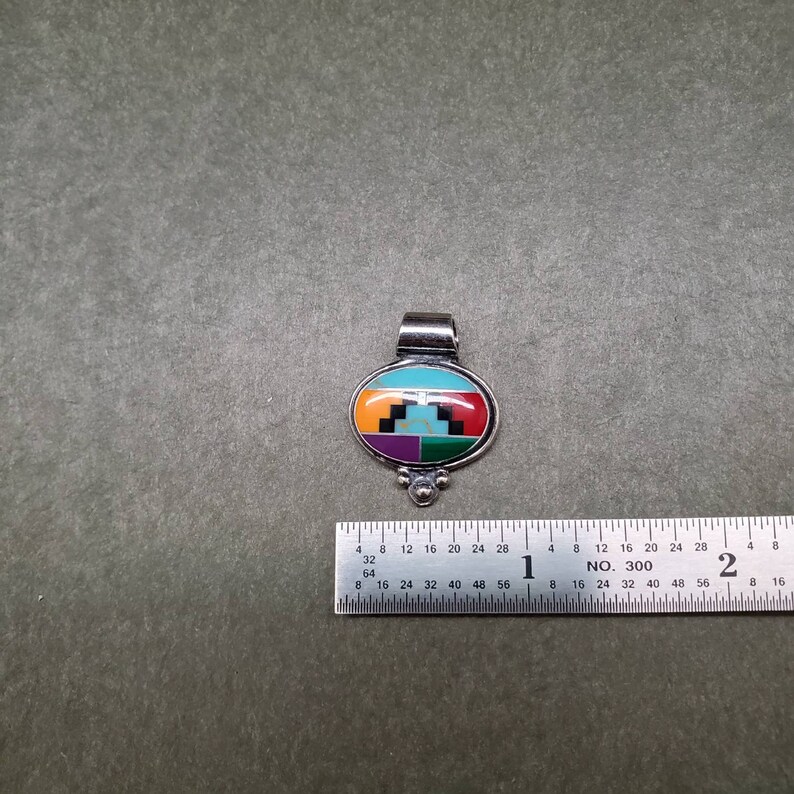 May include: A silver pendant with an oval-shaped inlay of turquoise, red, green, purple, and black stones. The pendant has a silver bail and three small silver beads at the bottom.