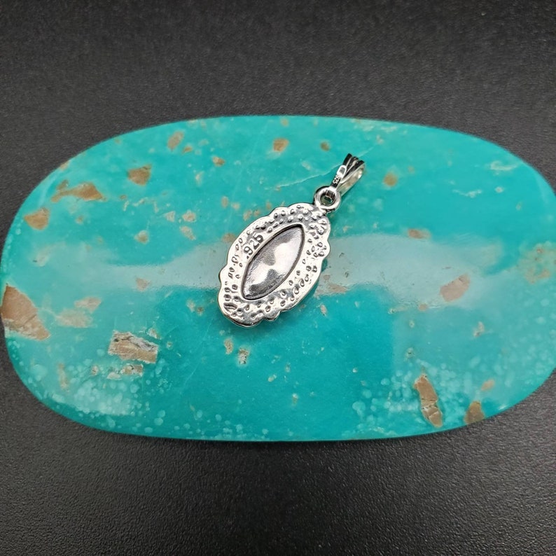 Peut inclure: Pendentif en argent avec un design ovale d&eacute;taill&eacute; et une pierre transparente au centre. Le pendentif est sur un fond turquoise.