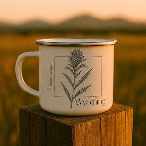 May include: A white enamel mug with a black rim and handle. The mug features a botanical illustration of a plant labeled "Castilleja coccinea" and the word "Wyoming" in a black font. The mug is set on a wooden post.
