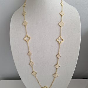 Op de afbeelding: Een goudkleurige ketting met klavervormige hangers met een crèmekleurig midden. De ketting wordt getoond op een witte mannequin, die de lengte en het ontwerp laat zien. De ketting is delicaat en de hangers zijn gelijkmatig verdeeld.