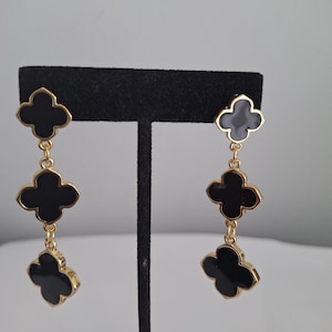 Peut inclure: Boucles d'oreilles pendantes dorées avec un motif de trèfle noir. Chaque boucle d'oreille comporte trois breloques en forme de trèfle reliées par de fines chaînes dorées. Les boucles d'oreilles sont présentées sur un support noir.