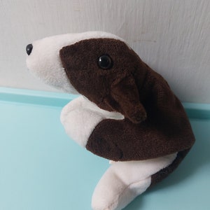 May include: A brown and white plush toy dog with black eyes. The dog is sitting on its hind legs with its front paws tucked in.