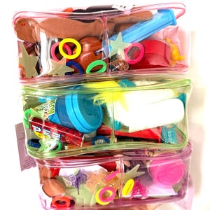 May include: Three clear, divided cosmetic bags stacked on top of each other, filled with colorful hair accessories and small toys. The bags are pink, green, and pink, and contain items like hair ties, star-shaped clips, and a Pez dispenser.