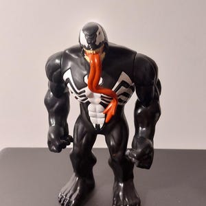 Peut inclure: Figurine d'action Venom noire et blanche avec une langue rouge. La figurine a une construction muscl&eacute;e et est pos&eacute;e dans une posture mena&ccedil;ante.