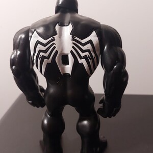 Peut inclure: Figurine d'action noire et blanche de Venom, un super-h&eacute;ros de Marvel Comics. La figurine est en position debout, les bras le long du corps. La figurine a une carrure large et muscl&eacute;e et un symbole d'araign&eacute;e blanc sur le dos.