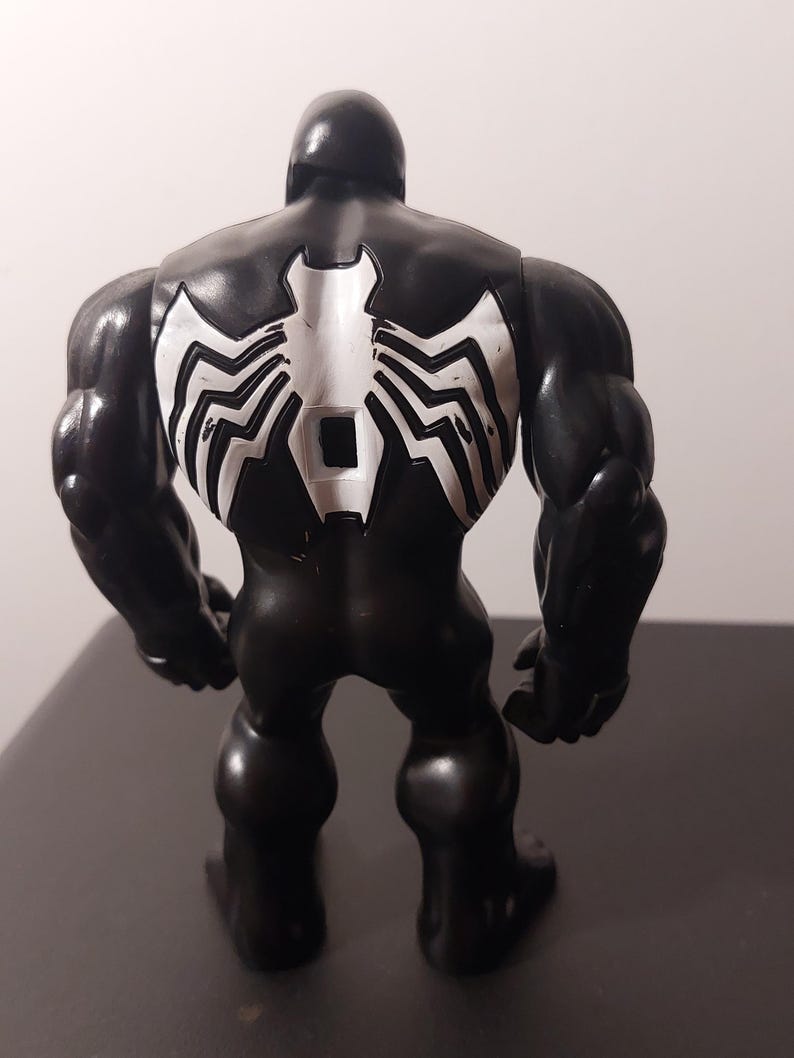 Peut inclure: Figurine d'action noire et blanche de Venom, un super-h&eacute;ros de Marvel Comics. La figurine est en position debout, les bras le long du corps. La figurine a une carrure large et muscl&eacute;e et un symbole d'araign&eacute;e blanc sur le dos.