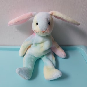 May include: A pastel tie-dye plush bunny rabbit toy with long floppy ears. The bunny is sitting with its legs crossed and has black button eyes.