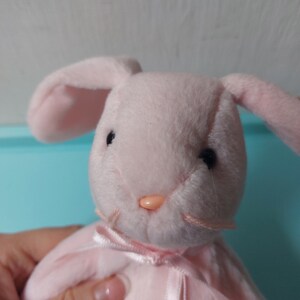 May include: A pink plush bunny with black eyes and a pink nose. The bunny is wearing a pink dress with a white ribbon.