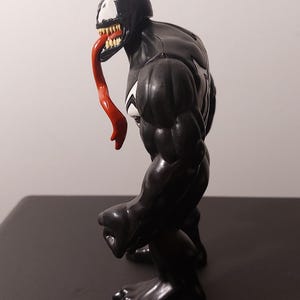 Peut inclure: Figurine d'action noire et blanche de Venom, un personnage de Marvel Comics. La figurine a une langue rouge et est pos&eacute;e debout, le bras droit pli&eacute; au coude.