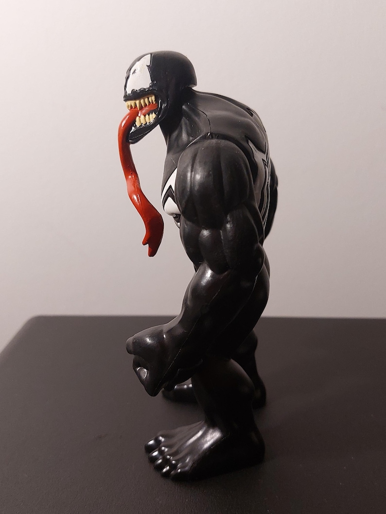 Peut inclure: Figurine d'action noire et blanche de Venom, un personnage de Marvel Comics. La figurine a une langue rouge et est pos&eacute;e debout, le bras droit pli&eacute; au coude.