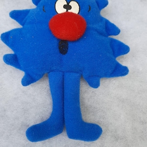 Puede incluir: Un peluche azul con forma de estrella con una nariz roja, ojos blancos y una boca negra. El juguete tiene dos piernas y pies azules. El juguete está hecho de tela suave.