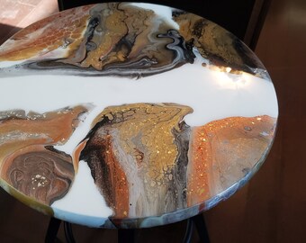 Puede incluir: Tapa de mesa redonda con una base de resina epoxi blanca y un patrón de remolino de colores marrón, negro, dorado y cobre. La resina epoxi tiene un acabado brillante y contiene purpurina dorada.