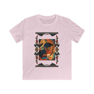 May include: A pale pink t-shirt with a framed illustration of a woman with dark hair and a floral design. The frame has geometric patterns in black, orange, and white. The shirt is made of a soft material.