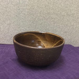 May include: A handmade, brown ceramic bowl with a textured exterior and a lighter brown interior. The bowl sits on a purple cloth, with a speckled gray background. The bowl's design features a simple, rustic aesthetic, suitable for serving or display.