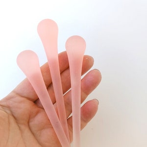 May include: Four pale pink glass stir sticks with rounded tops. The sticks are held in a hand against a white background. The glass has a frosted appearance.