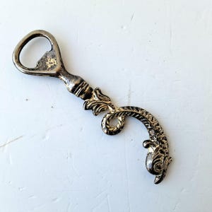 May include: A vintage silver-toned bottle opener with an ornate, fish-shaped design. The opener features a classic bottle cap removal end and a decorative handle with floral and scroll details. The metal has a slightly aged appearance.
