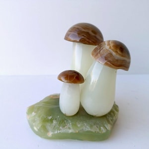 May include: A decorative sculpture of three mushroom figures with brown and white caps and stems. The mushrooms are set on a green, textured base. The sculpture is made of a glossy material.